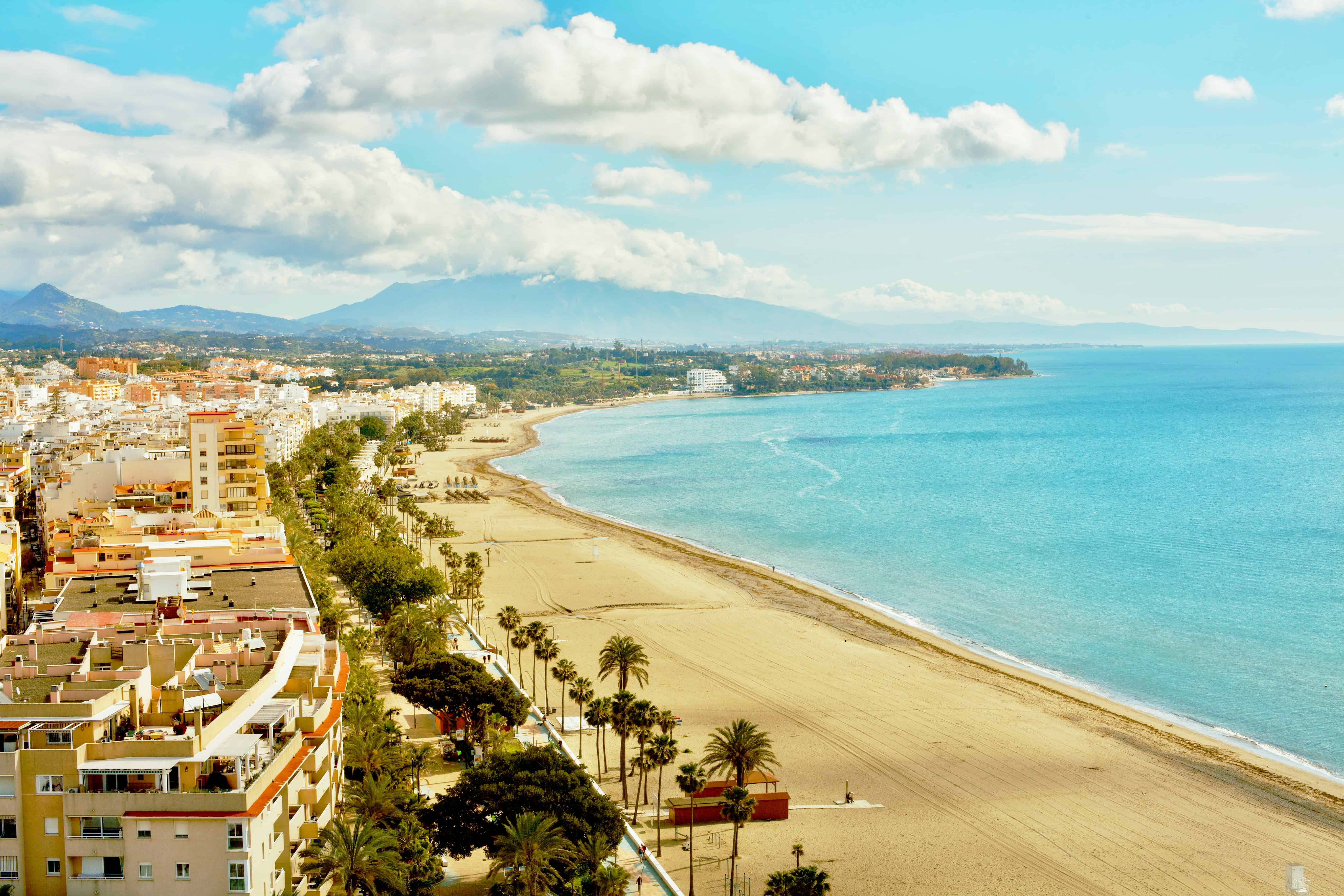 Luchtfoto van de brede zandstranden en boulevard van Estepona aan de Costa del Sol, met de Middellandse Zee en bergen op de achtergrond.